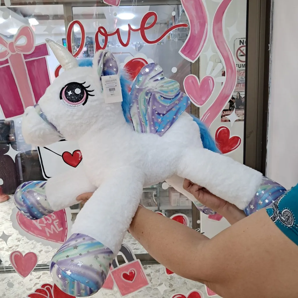 Peluche de unicornio acostado blanco con detalles galácticos tornasol en mostrador de tienda Valeland Barbosa