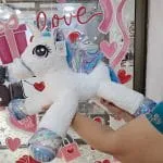 Peluche de unicornio acostado blanco con detalles galácticos tornasol en mostrador de tienda Valeland Barbosa