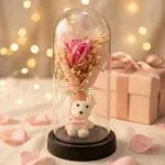 Cúpula de rosa eterna rosada con tierno osito blanco encendida con luces LED junto a caja de regalo rosa y pétalos con fondo bokeh