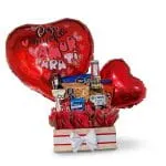 Ancheta de madera con cerveza Coronita Extra, variedad de snacks, chocolates y globos de corazón metalizados rojos sobre fondo blanco